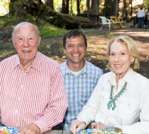 Three people sitting side by side smiling with a redwood forest and other people in the background