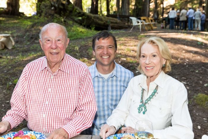 Three people sitting side by side smiling with a redwood forest and other people in the background