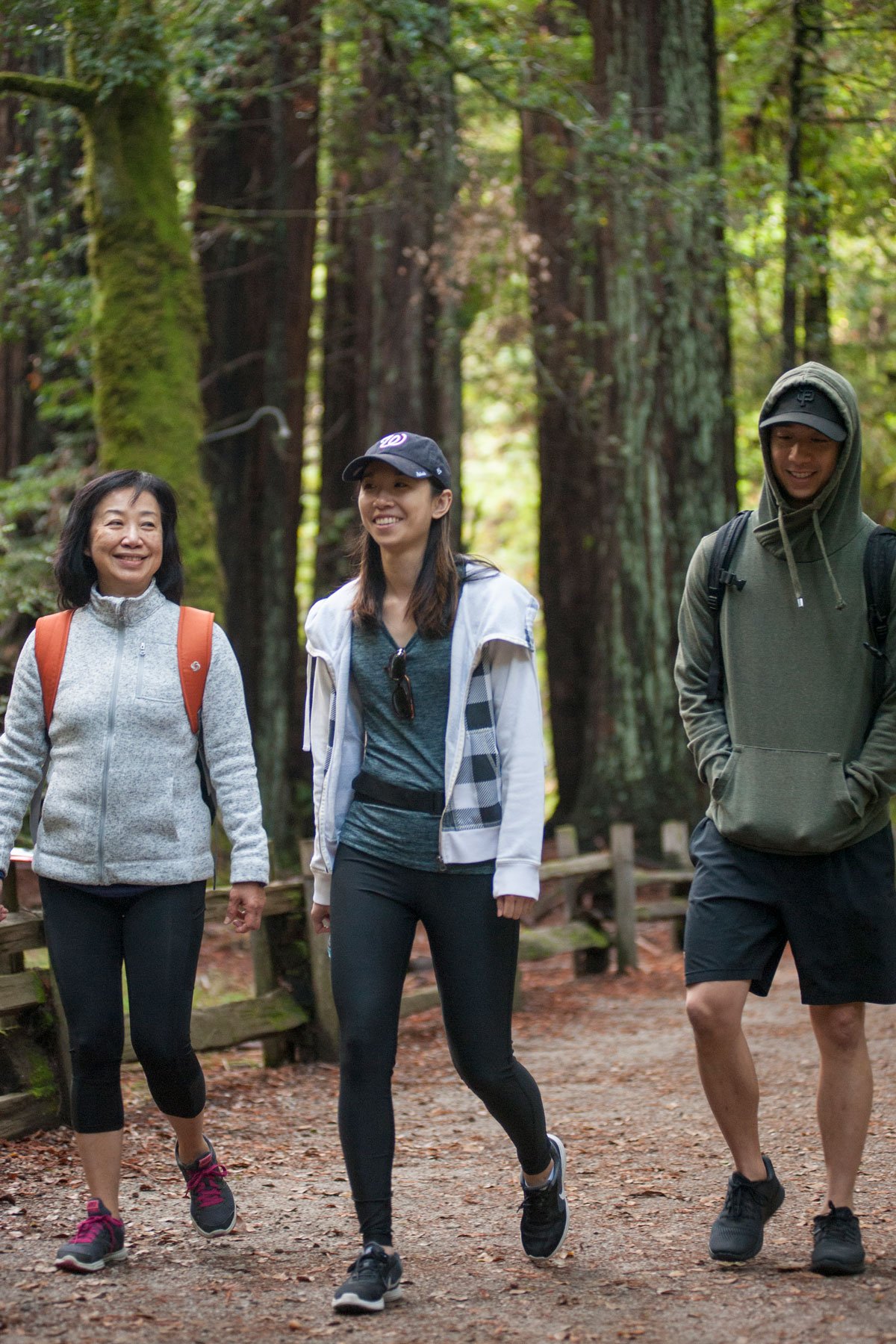 An older woman, a young woman, and a young man, all of asian descent, walk through the redwoods with smiles on their faces.
