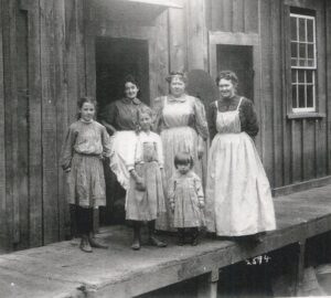 Three women in aprons stand with three young girls in dresses in front of a wooden structure, circa late 1800s/early 1900s.