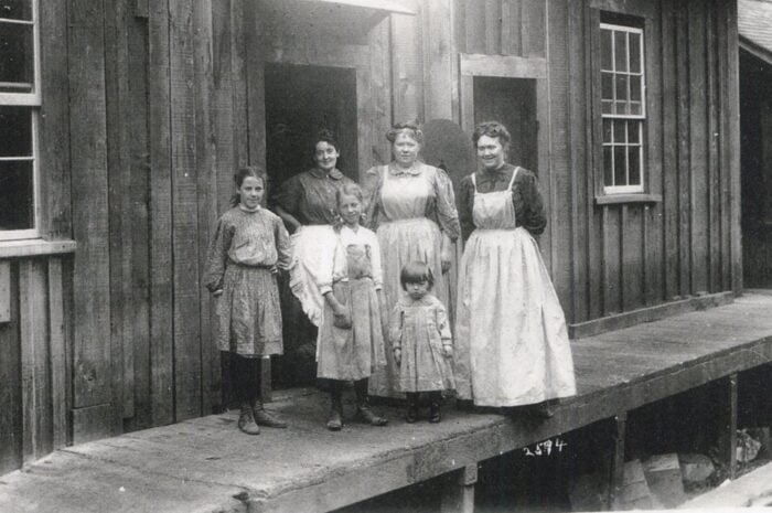 Three women in aprons stand with three young girls in dresses in front of a wooden structure, circa late 1800s/early 1900s.