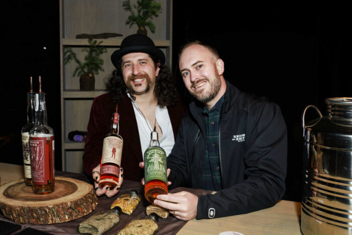 two men smile at the camera while holding up bottles of whiskey