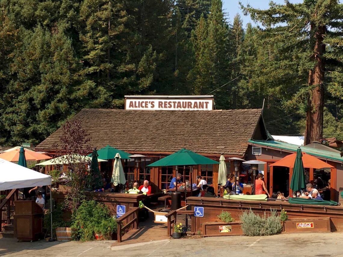 A restaurant with an umbrella-shaded porch and sign reading Alice's Restaurant with tall redwoods in the background