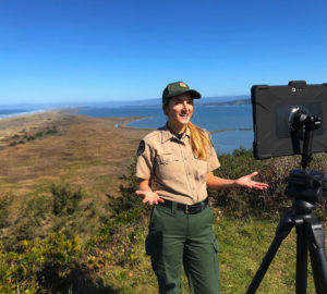 A female California State Parks naturalist interpreter in park ranger fatigues gives her presentation before a tablet on a tripod on which she is livestreaming a virtual program to Facebook.