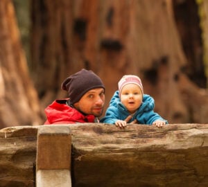 A man and a baby, both wearing knitted caps, sitting on a bench in a giant sequoia forest