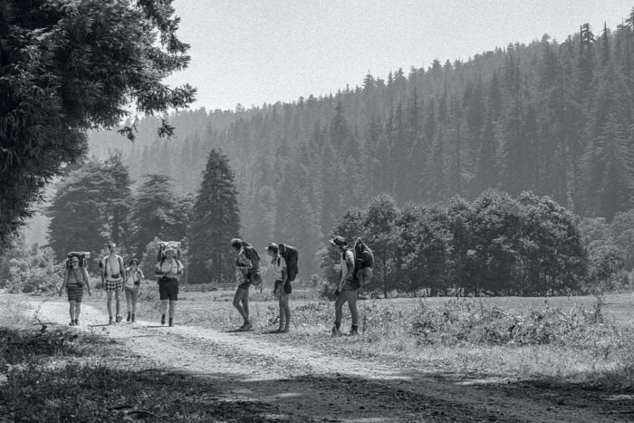 Black-and-white photo of seven backpackers hiking on a flat, open trail surrounded by conifer forests on a mountain.