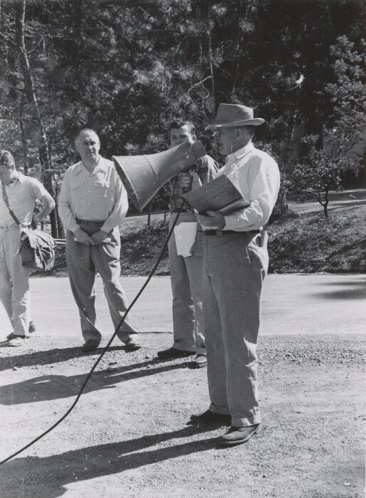 Black and white photo of a man holding a clipboard, speaking through a large megaphone to other assembled men