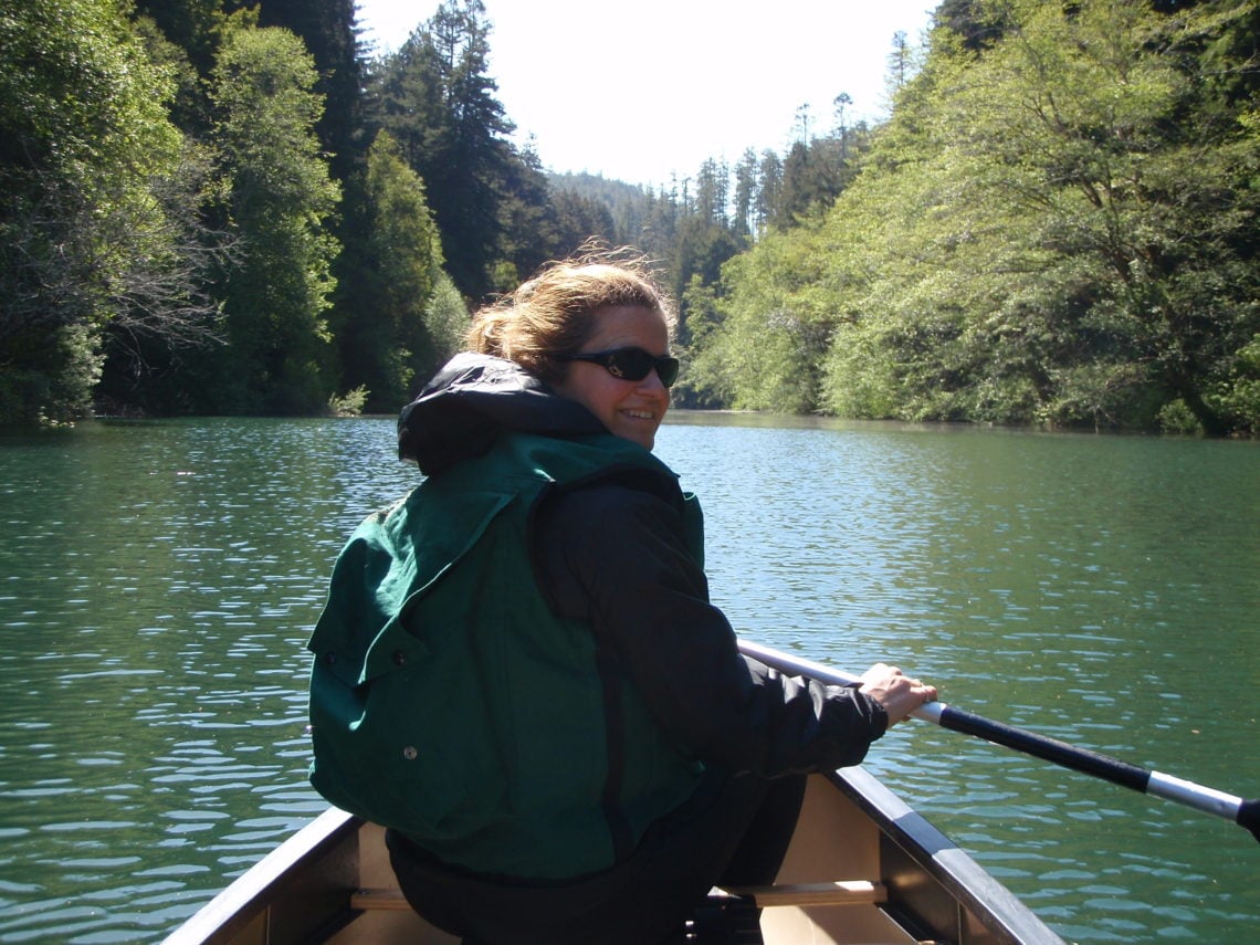 In the foreground, a smiling person paddles a canoe down a river on a sunny day. Trees line the shore.
