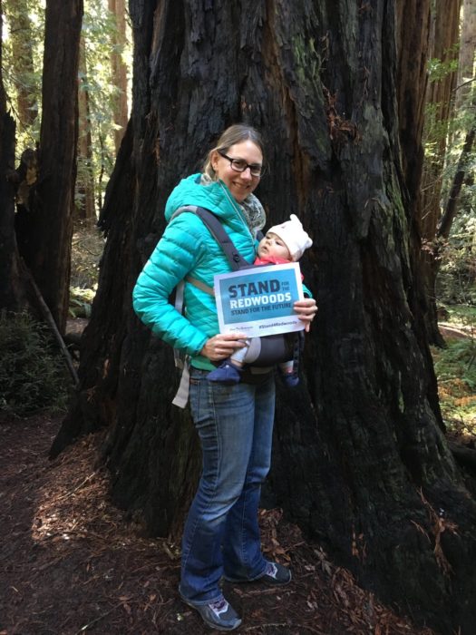 A woman wearing a turquoise puffer jacket and eyeglasses, holding a baby in front of a coast redwood tree.