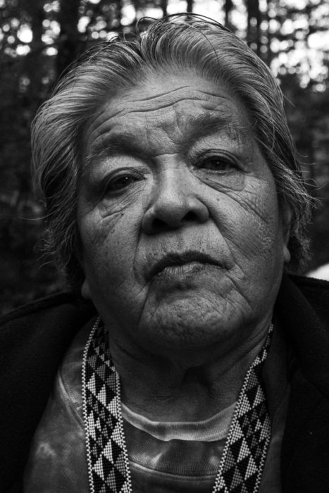 A black and white portrait of an elderly native american indigenous woman wearing a beaded necklace with a serious expression.