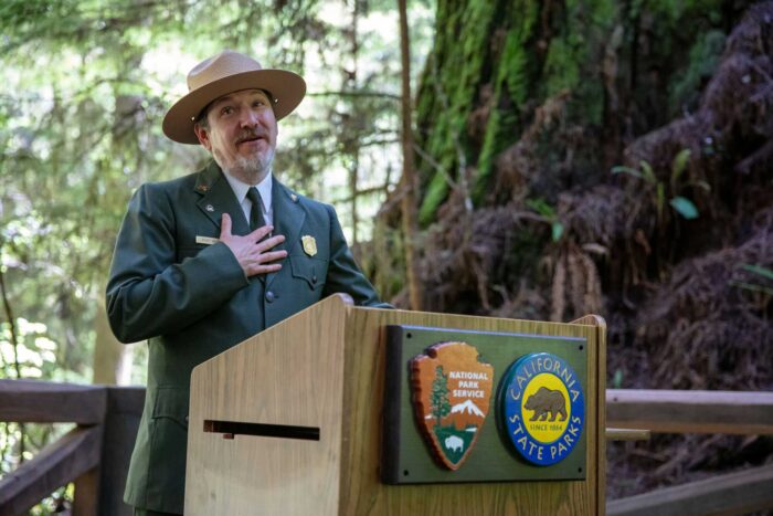 man in green jacket and round hat stands at podium in front of redwood tree.