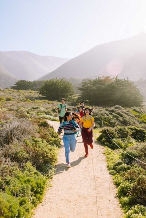 A group of 5 young people of color on the trail in Garrapata State Park, surrounded by coastal brush on a sunny day with mist in the air. Two women in the front are running and smiling.