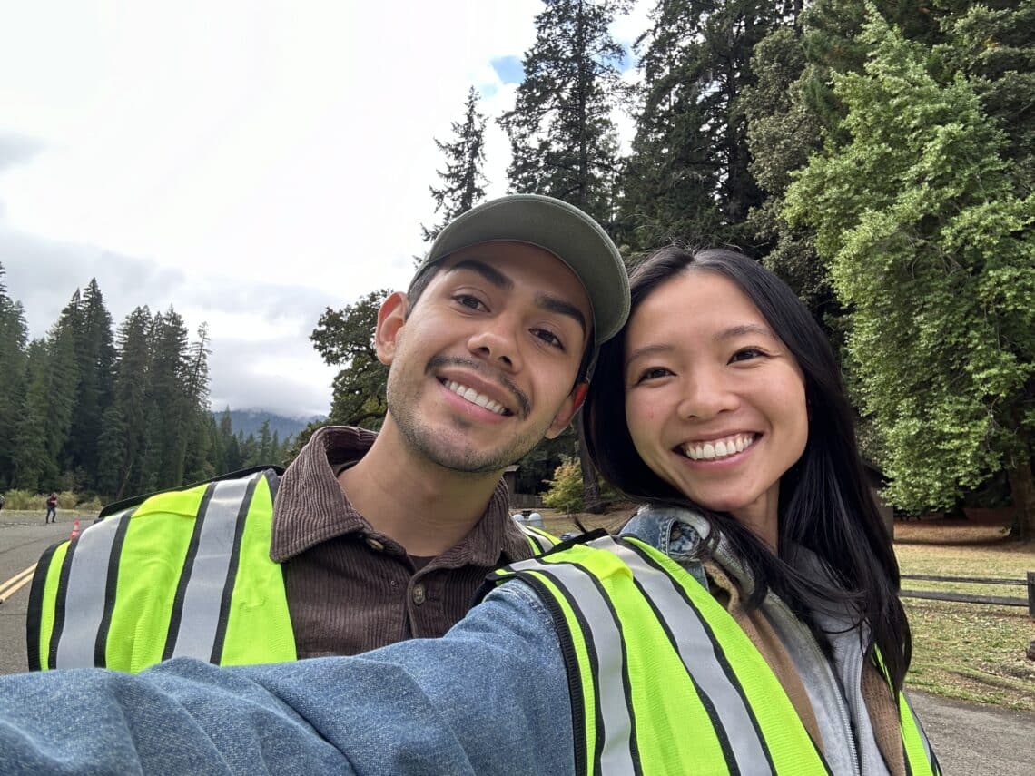 Two young people pose for a selfie in front of tall redwood trees.