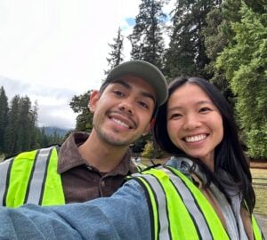 Two young people pose for a selfie in front of tall redwood trees.