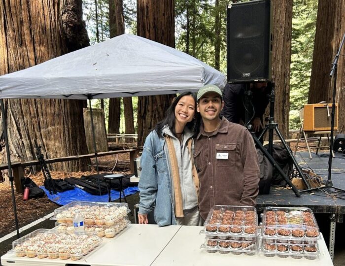 Two friends stand behind a table stacked with vanilla and chocolate cupcakes.