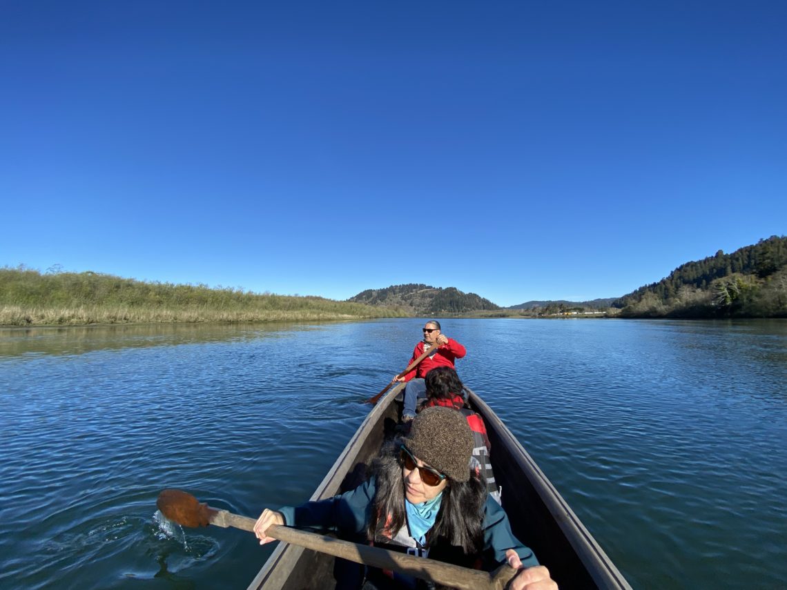 Three people sit in a dugout canoe on a sunny day. The people in the foreground and background paddle down a river with large wooden paddles.