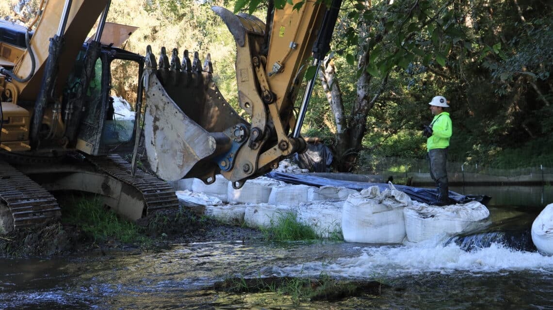 Construction equipment removes a dam from a stream while a worker looks on