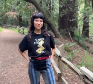 A woman with dark hair wearing a black t shirt and blue jeans stands on a trail in a redwood forest