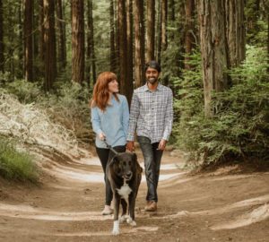 A white woman with red hair and bangs and an Indian man walk their large dog in Joaquin Miller, holding hands and smiling.