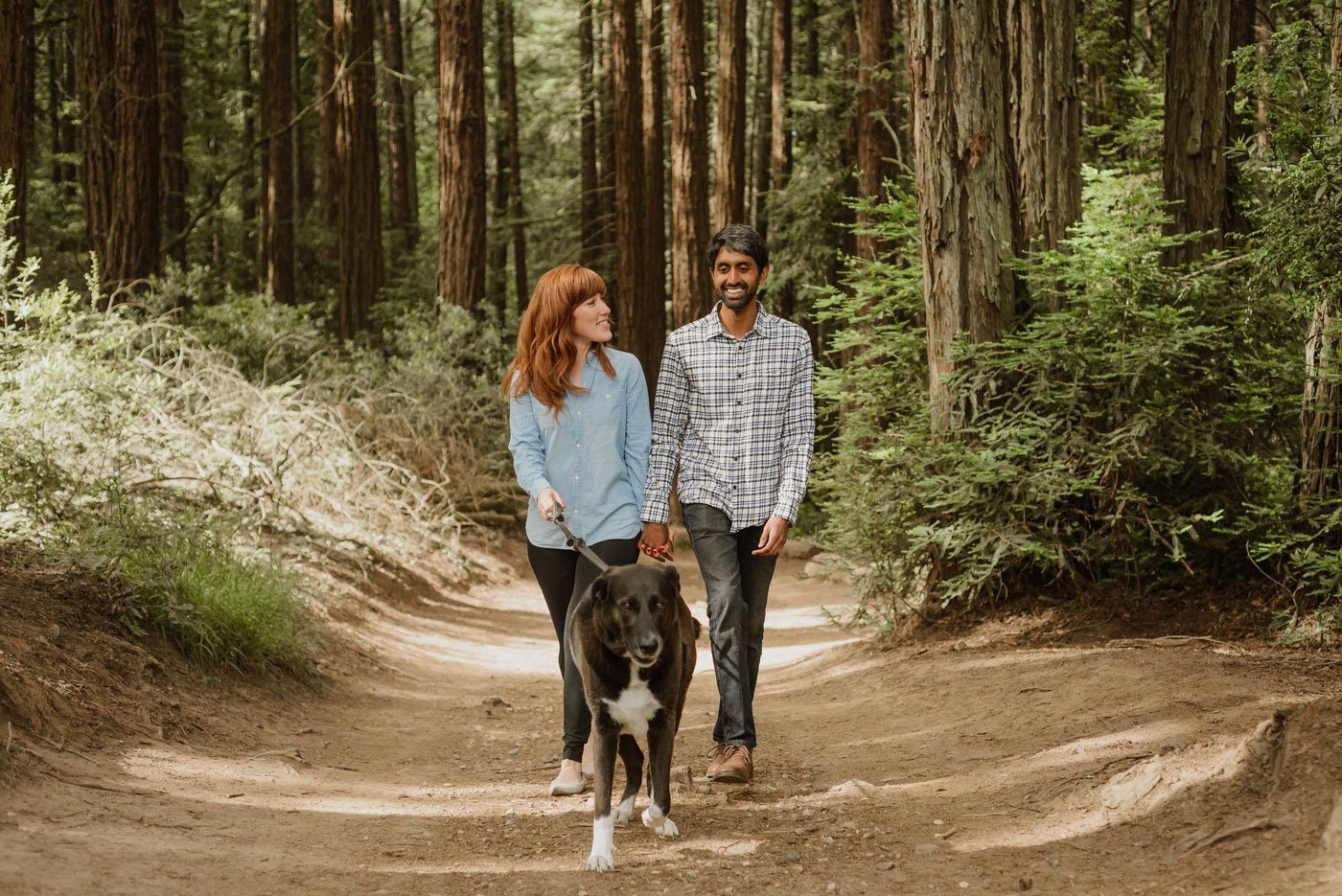 A white woman with red hair and bangs and an Indian man walk their large dog in Joaquin Miller, holding hands and smiling.