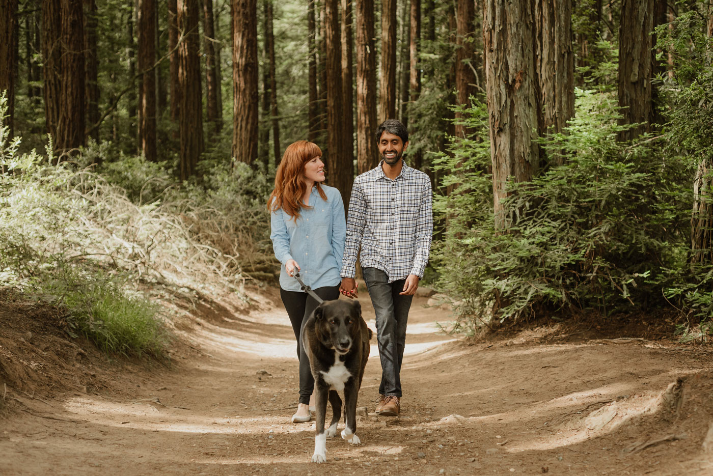 A white woman with red hair and bangs and an Indian man walk their large dog in Joaquin Miller, holding hands and smiling.