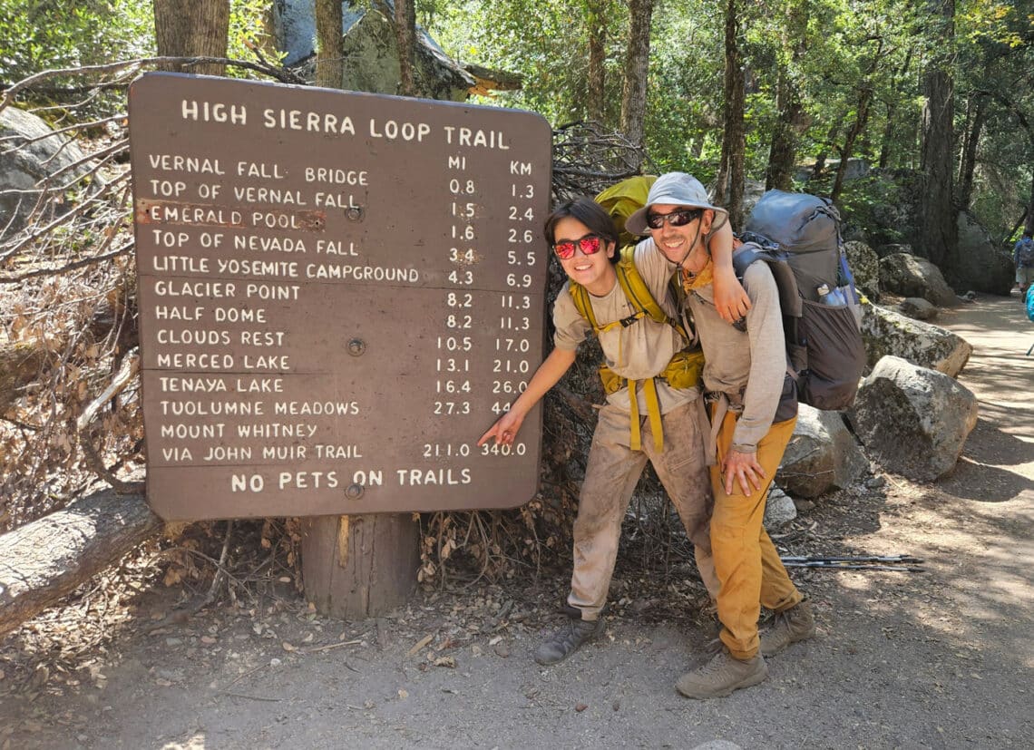 A teen boy and his father in muddy earth tone clothes, hiking backpacks, and sunglasses, smiling and pointing at a large park sign titled High Sierra Loop Trail. The teenager is pointing to the 211-mile long John Muir Trail route.