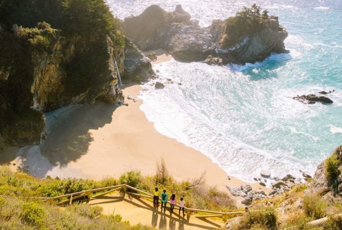 Four people wearing brightly colored shirts look out past McWay Falls towards the bright blue sea.