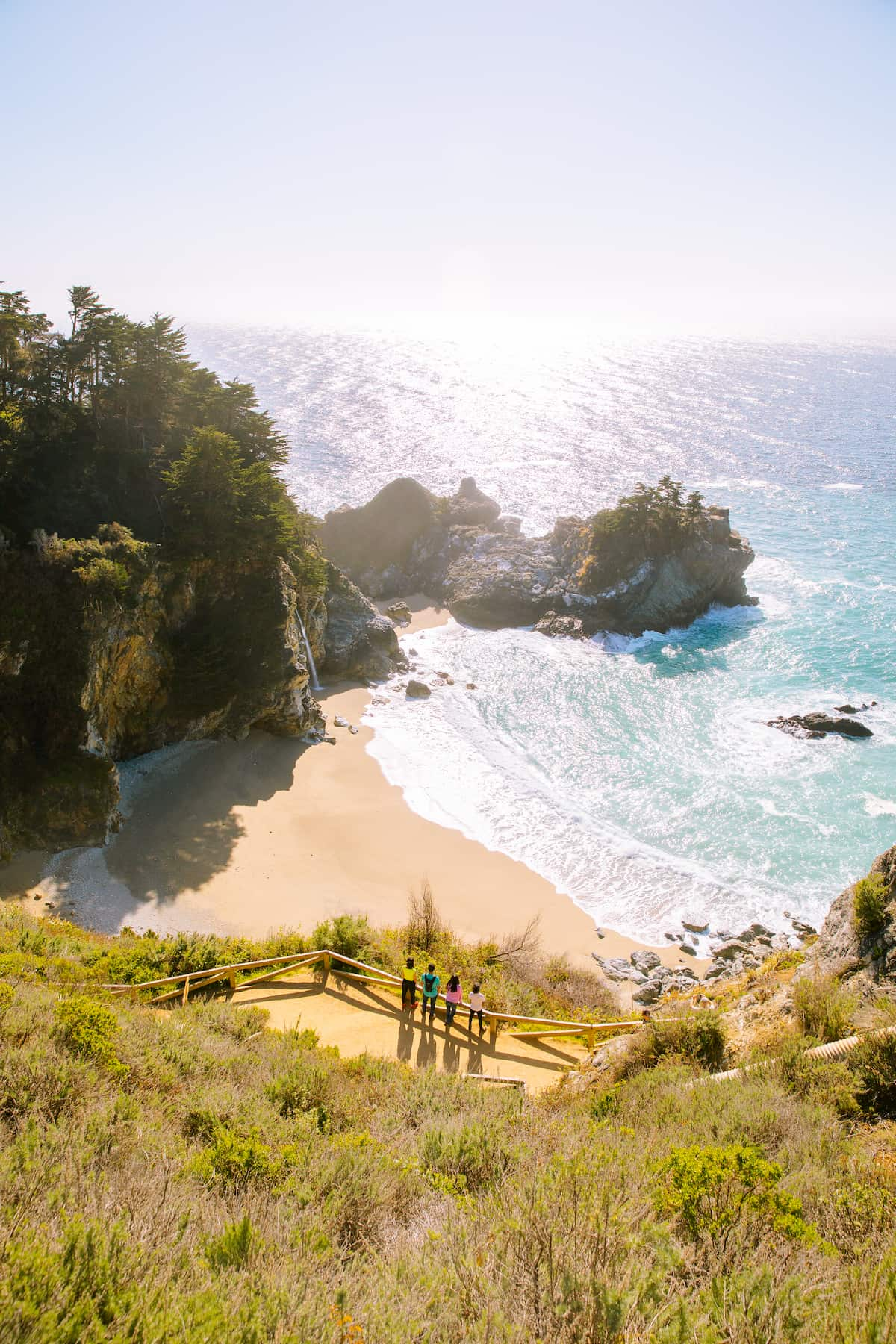 Four people wearing brightly colored shirts look out past McWay Falls towards the bright blue sea.