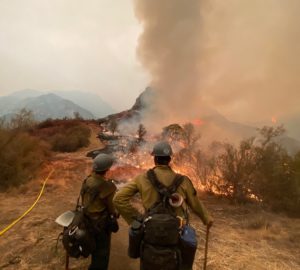 Two firegfighters stand on a ridge with fire and smoke