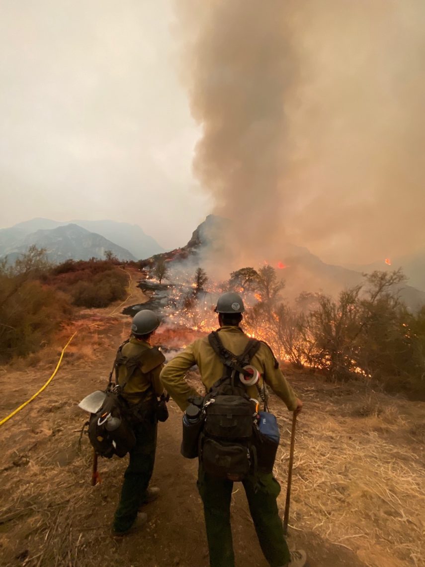 Two firegfighters stand on a ridge with fire and smoke