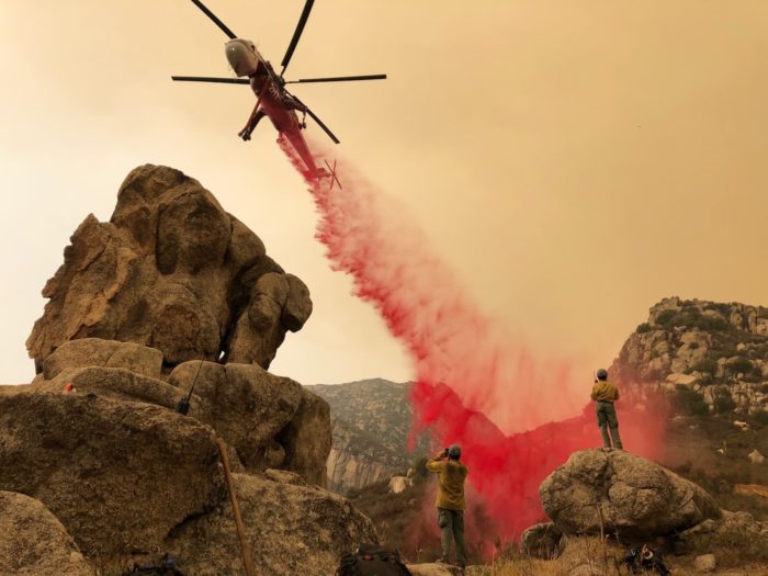 A helicopter drops pink fire retardant on a mountain as two firefighters stand below