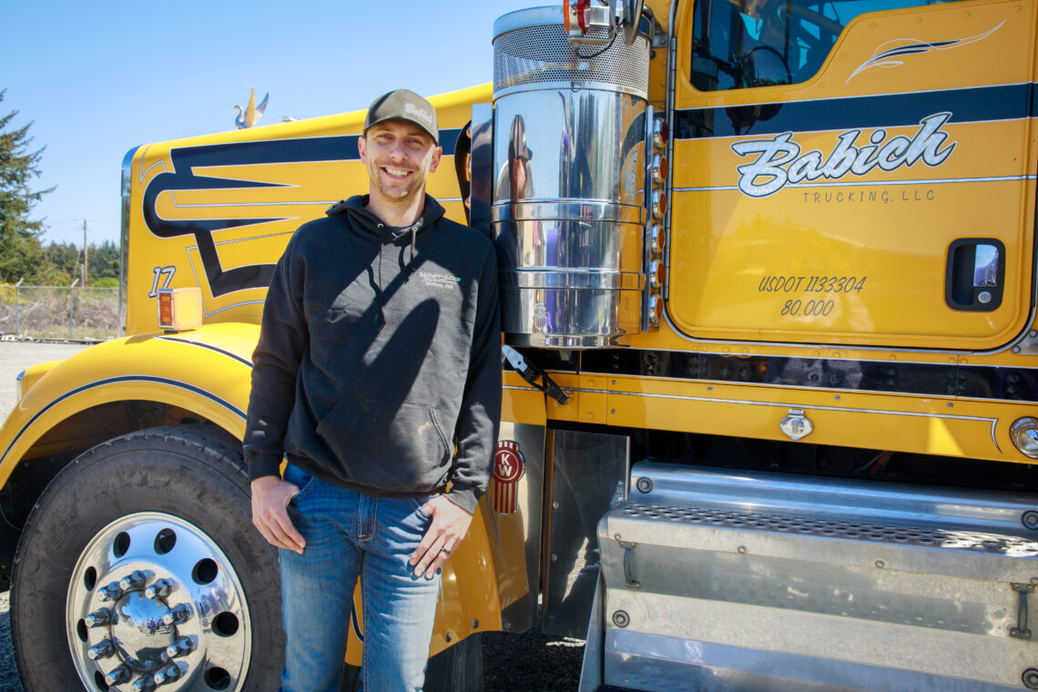 A young man wearing jeans, a black hoodie and a ball cap smiles at the camera. He leans against a bright yellow big rig with the word Babich on the side.