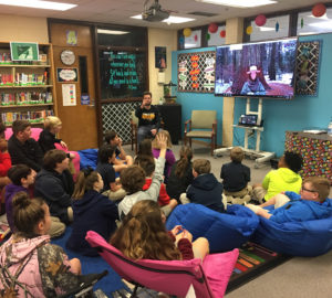 A classroom of school children gather around a screen to watch a video. 