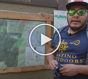 A male representative of Latino Outdoors stands before an interpretive trailhead sign depicting a map of Nadler Grove in Sierra National Forest.