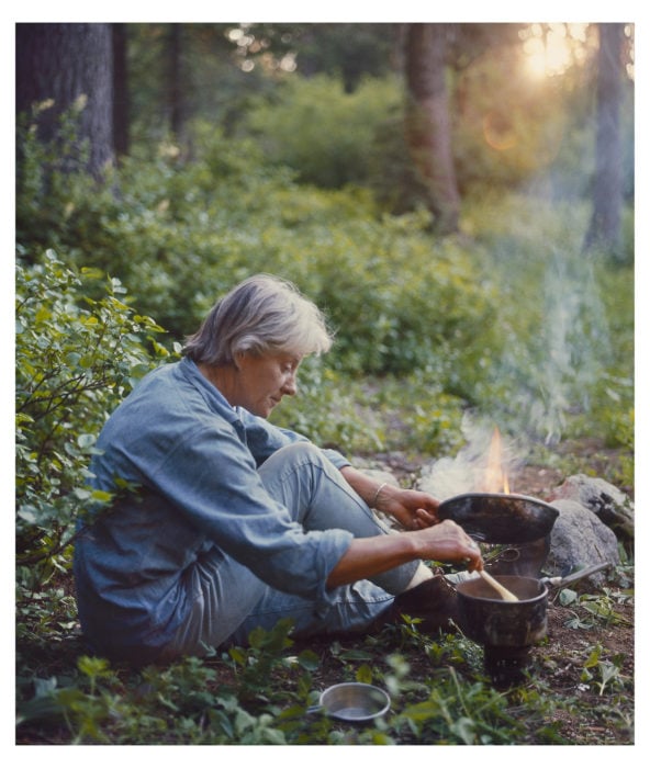 A grey-haired woman sits and cooks on a camp stove, surrounded by greenery