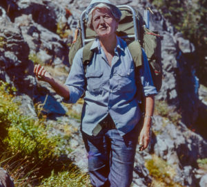 A grey-haired woman backpacking, with greenery and boulders in the background.