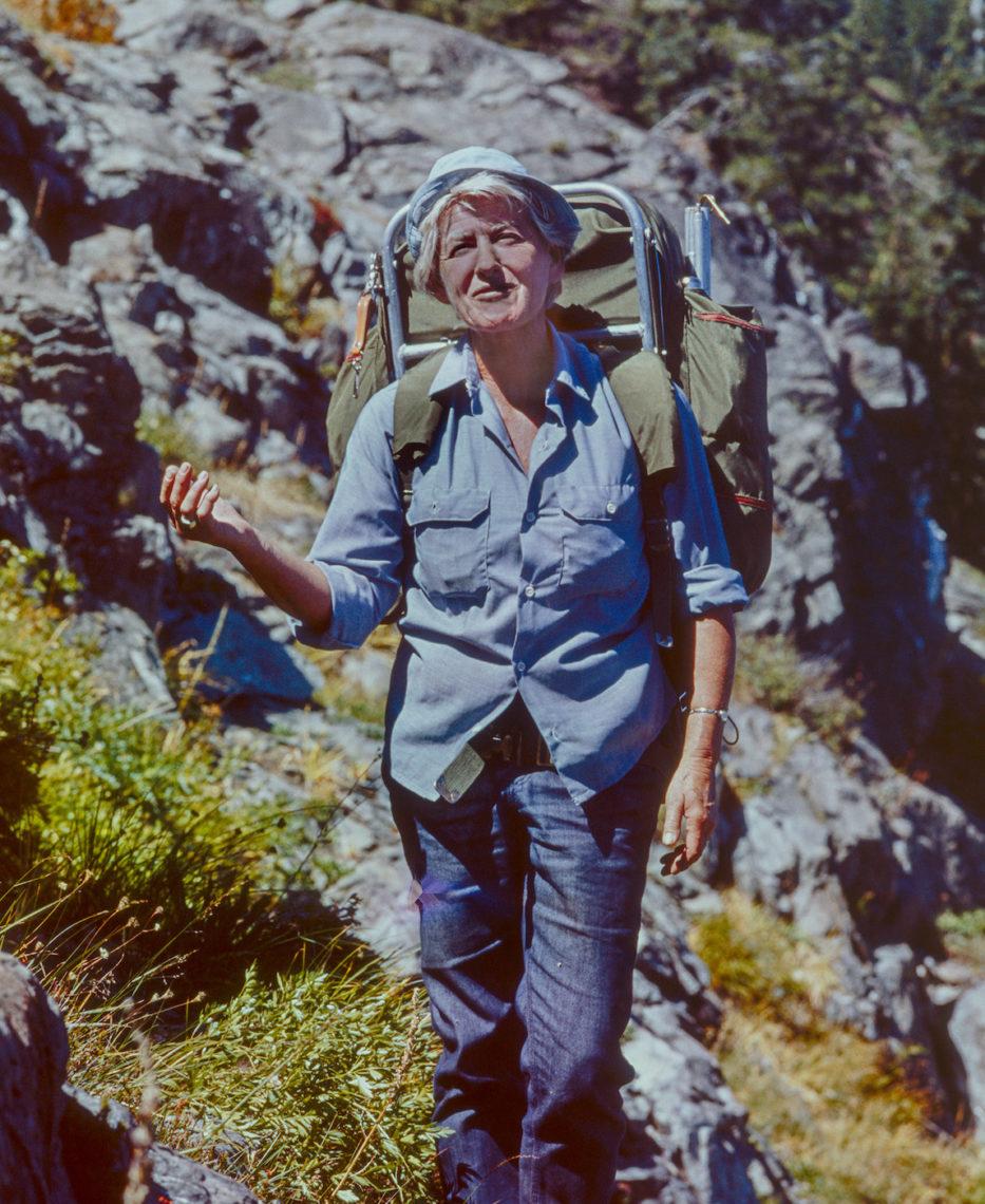 A grey-haired woman backpacking, with greenery and boulders in the background.