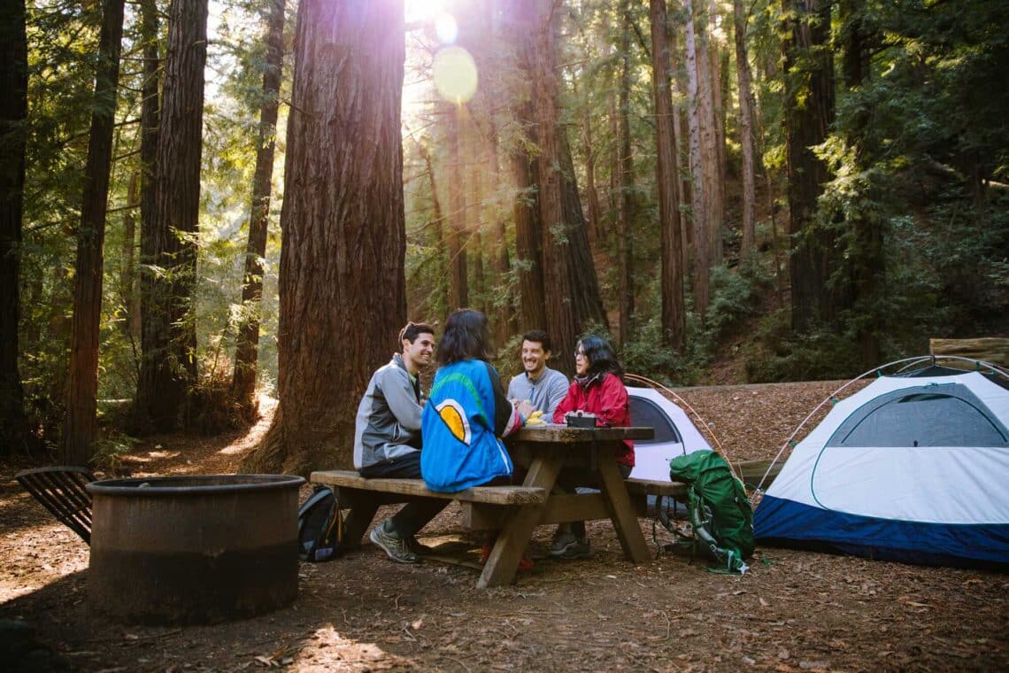 Four people sit at a picnic table with two camping tents and redwood trees in the background