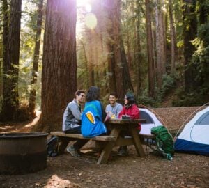 Four people sit at a picnic table with two camping tents and redwood trees in the background