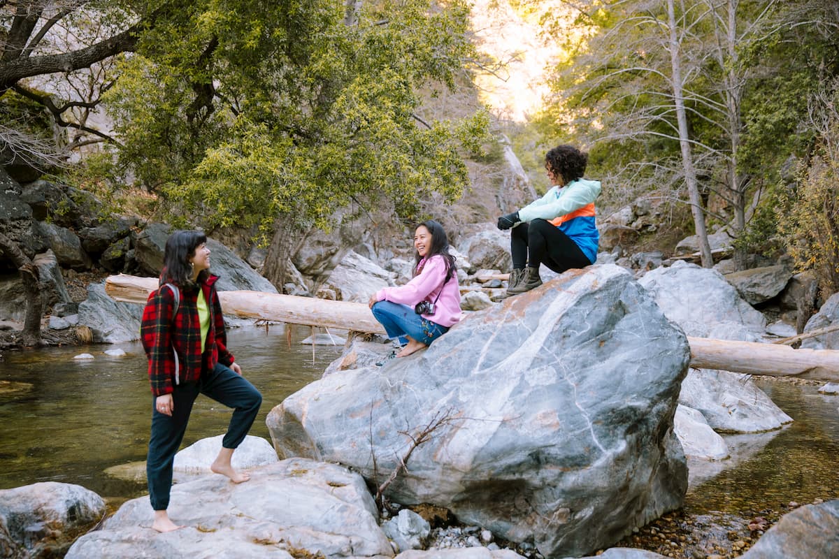 Three women of color smile and laugh with each other as they sit on a boulder in a creek in a redwood forest.