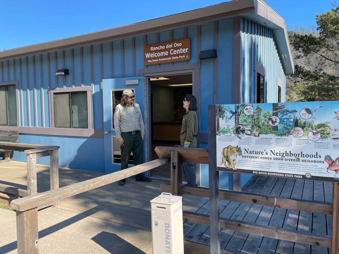 A ranger standing in front of the new Rancho del Oso Welcome Center (a blue building with interpretive signage in front.)