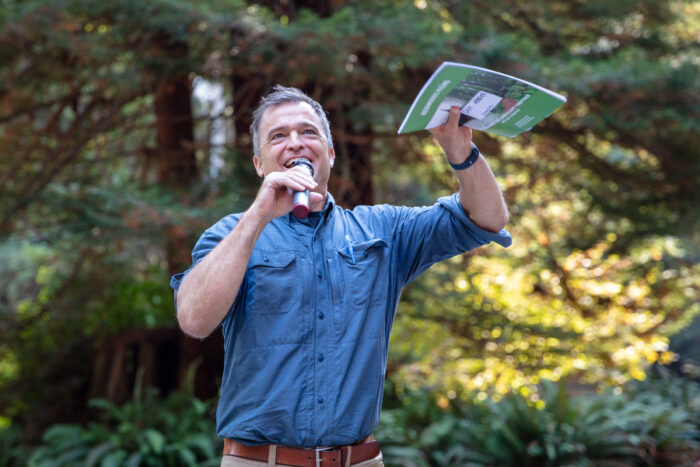 A man in a blue shirt with a microphone smiles as he addresses the crowd