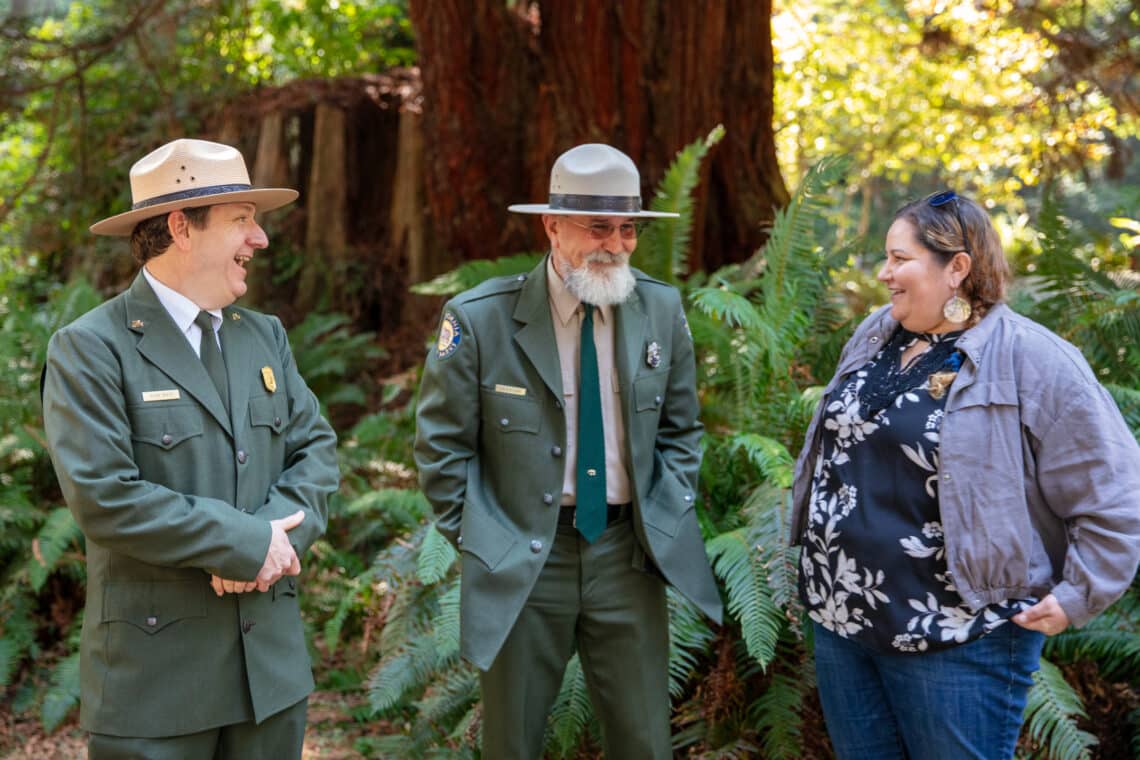 Two men in olive green state park uniforms with ranger hats smile as they speak to a woman in civilian clothes
