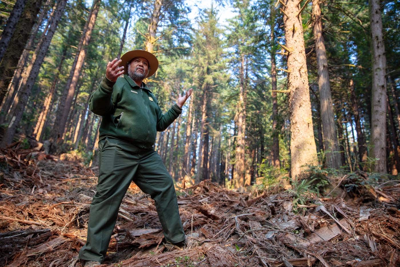 A man of color in olive jacket and pants standing in woody debris in a forested area.