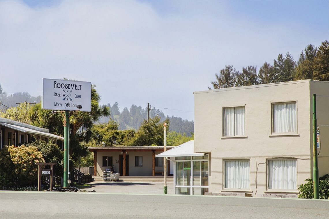 A simple cream-colored building with a sign reading Roosevelt Base Camp Motel and Lodge, with a redwood forest in the background