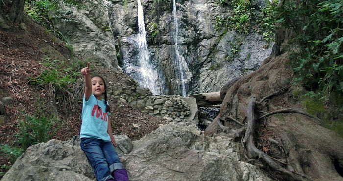 A kid sits on a large rock in front of a small waterfall over a rock wall