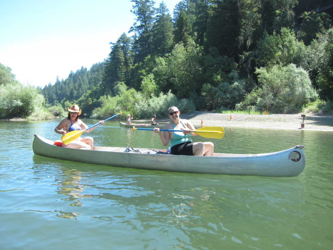 In the foreground and background, people paddle two canoes on a river on a warm, sunny day. Trees and brush line the shores.