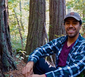 A hispanic man of Mexican heritage in a baseball cap, blue flannel, purple shirt and dark pants is sitting in a redwood grove and smiling.