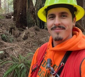 A close up selfie of a young man of Yurok heritage with a goatee wearing a yellow construction hat, orange Redwoods Rising hoodie, red vest with a camelback attachment standing in a redwoods forest.