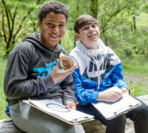 Two smiling boys sit on a log holding clipboards. One holds a cross-section of a tree.